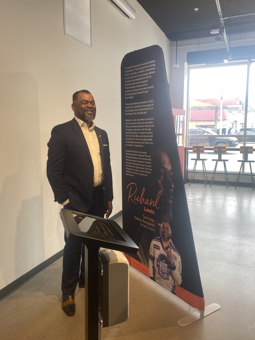 Richard Lewis standing next to a banner that tells the story of his two kidney transplants.