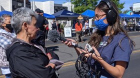 Una reportera con cubreboca, micrófono, grabadora y audífonos, entrevista a una señora en medio de un festival callejero.