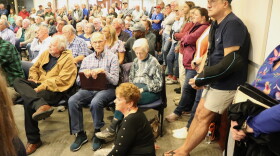 Hundreds of residents sit and watch a Leland mayor and council meeting in the chambers. Many are standing along the walls. One elderly woman sits on the floor in the center of the frame. An elderly man seated in a chair to her left stares at the camera.