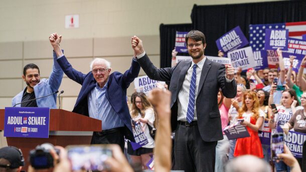 Three men stand holding hands in the air in celebration in front of a crowd of people.