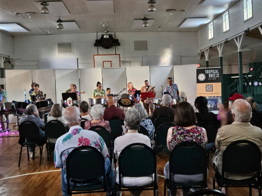 A steel band performs inside a community hall while an audience watches from rows of chairs.
