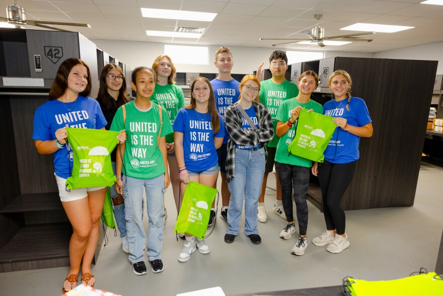 A group of ten teen volunteers in blue and green "United Is the Way" T-shirts stand together in a locker room, several holding up green United Way drawstring bags with a parachute logo reading "United Is the Way to Bright Futures.