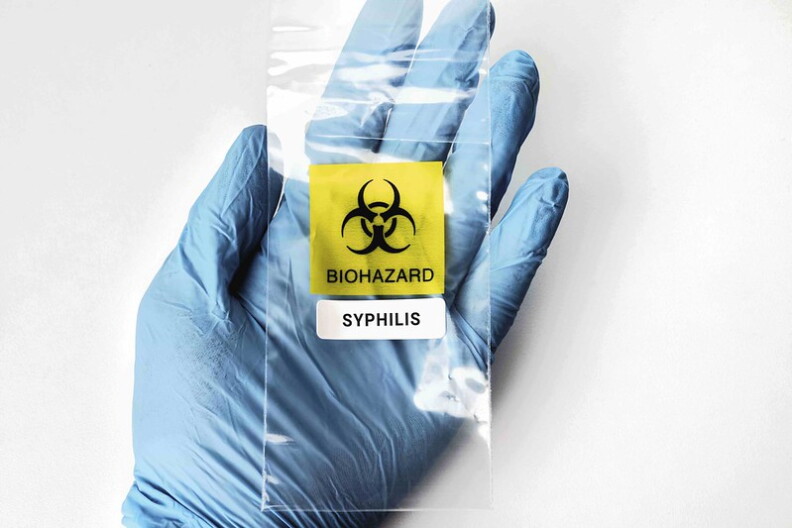 A hand wearing a blue medical glove holds a plastic bag with a biohazard symbol and a label of "syphilis."