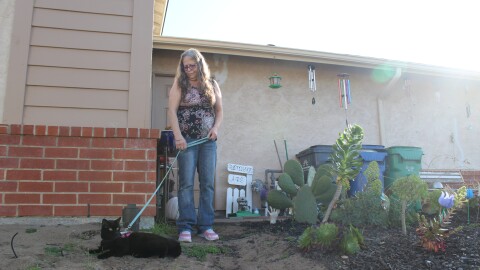 A woman wearing jeans and a patterned tank top holds a black cat on a leash in a patch of sandy dirt in front of a beige house with brick trim.
