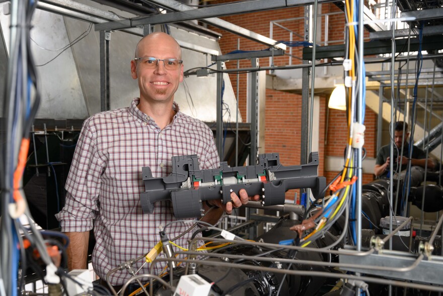 A man smiles while holding up a piece of machinery, while surrounded by wires in some sort of lab or factory