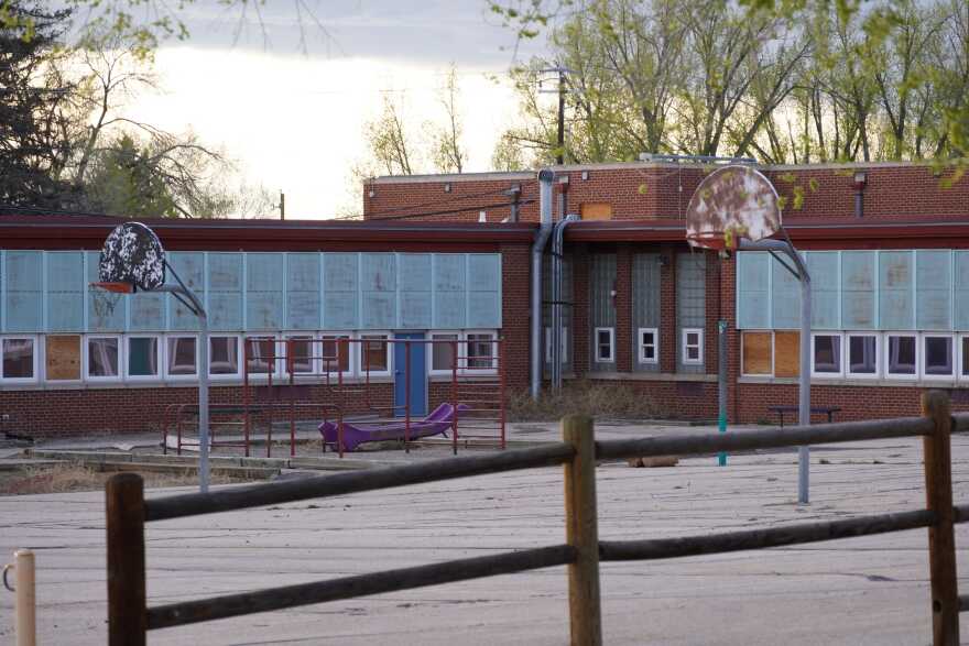 A dilapidated school building with boarded up windows surrounds a dirty paved playground, with dirty basketball hoops.