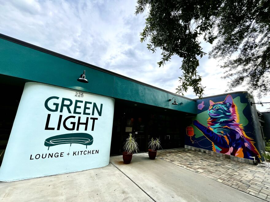 Exterior of a building says Green Light Lounge + Kitchen on a white wall with a mural of a purple cat to the right