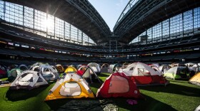  Several colorful camping tents are spread across the outfield grass of a professional baseball stadium as the sun shines through glass paneling.