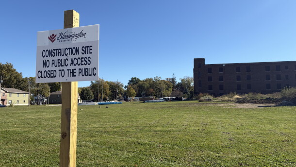 in the foreground, a white sign on a wooden post. in the background, an open field and brown brick building