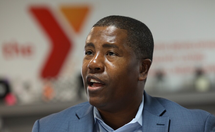 A smiling man wearing a light blue collared shirt under a blue-grey suitcoat. The orange and red YMCA logo is in the background.