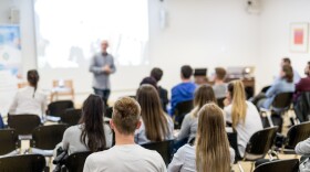 A speaker lectures in a university lecture hall as we see the backs of seated students listening