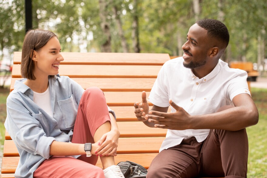 Happy young intercultural couple in casualwear sitting on wooden bench in park or square, chatting and enjoying summer day at leisure