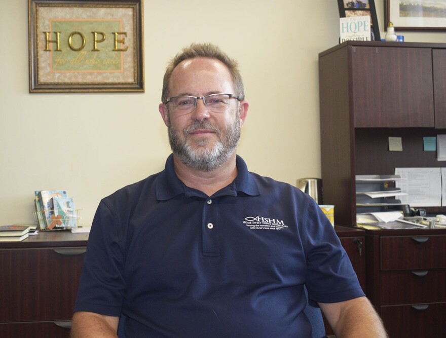 Man in a blue buttoned-down shirt seated in an office with a framed sign that reads 'HOPE' on the wall behind him