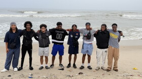 A group of young men with their arms around each other, standing on sand in front of a body of water