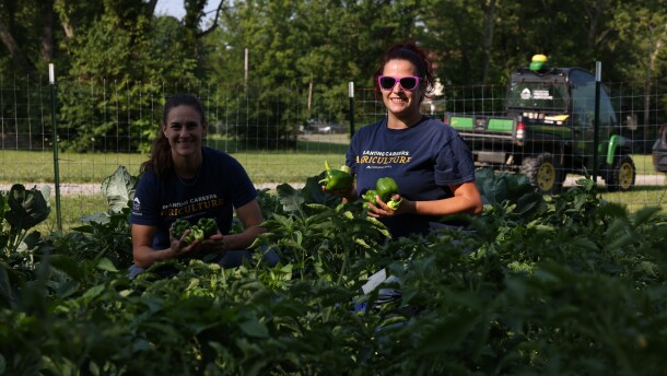 Two women kneeling in a garden of crops, holding up vegetables harvested on-site