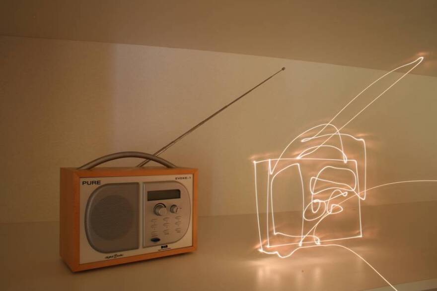 A small portable radio with an extended wire antenna sitting next to a glowing, illuminated wire sculpture of a radio on a table
