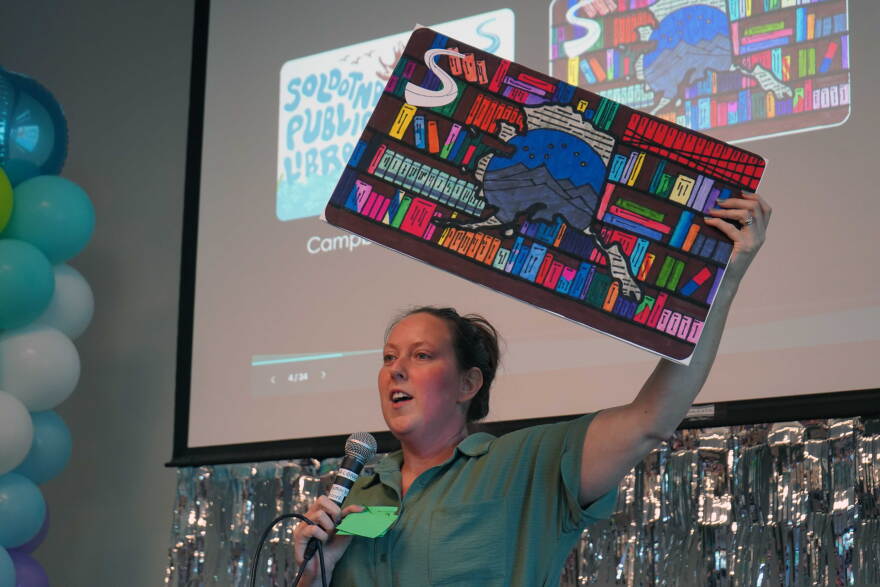 Kim McMilin holds aloft an enlarged prop version of one of three new limited edition library card designs unveiled Tuesday, Sept. 1, 2026 at the Soldotna Public Library in Soldotna, Alaska.