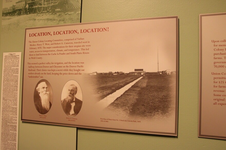 A placard inside a museum reads, "Location, Location, Location!" The picture shows some of Greeley's key people in founding the city as well as an old, black and white photo that shows some buildings in the rural area and a water canal running through it. 