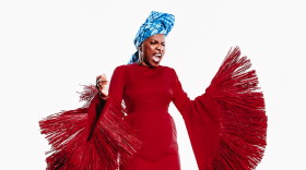 A woman in a dramatic, tassled. floor-length red dress and blue head turban, singing forcefully