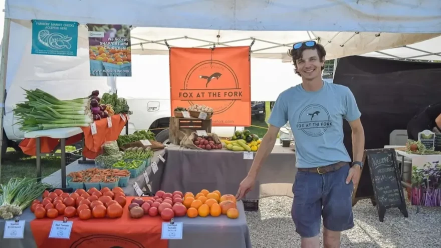 Man standing in a farmers market tent next to fresh produce.