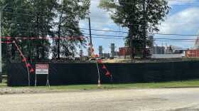 A black fence with a sign that reads "Private Property -- No Trespassing" stands in front of a property that contains smoke stacks, shipping containers and other industrial equipment. 