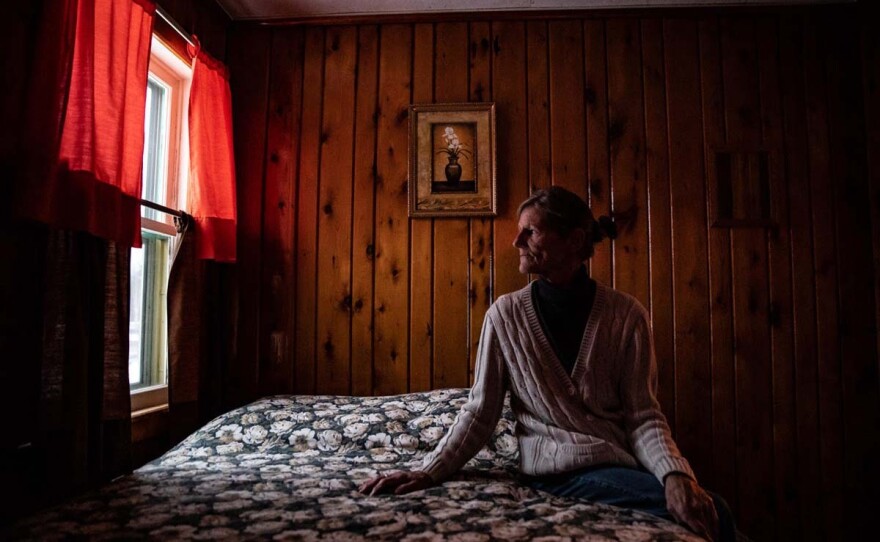 Robin Saunders, an older white woman, sits on a bed in a room with wood panelling and stares out a window