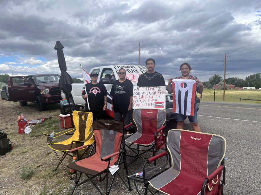 Four people hold protest signs and flags in front of a truck with more signs on it. Lawn chairs are set up in front of them.