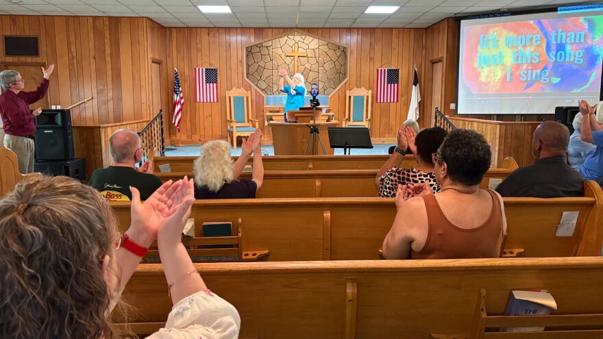 People in pews raise their joined hands as they look to a minister behind a pulpit.