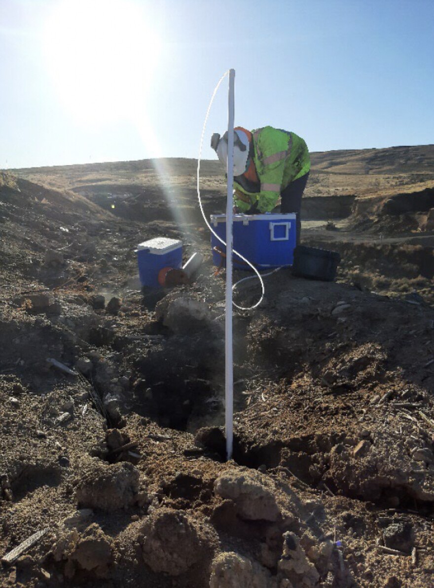 A worker in a yellow safety jacket reaches into a blue cooler. A white pole is sticking out of the ground in front of the worker.