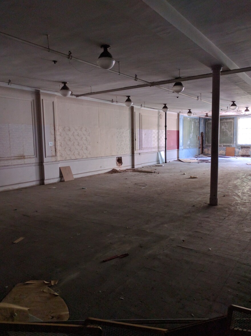 A view of a long, mostly empty, former commercial space with windows at one end. The ceiling has a series of older-looking small spherical light fixtures. The wall has wallpaper in some places, paint in others and might have been stripped at the far end.