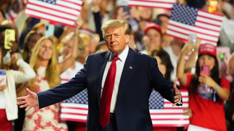 President Donald Trump stands with his arms outstretched. A crowd behind him waves American flags.