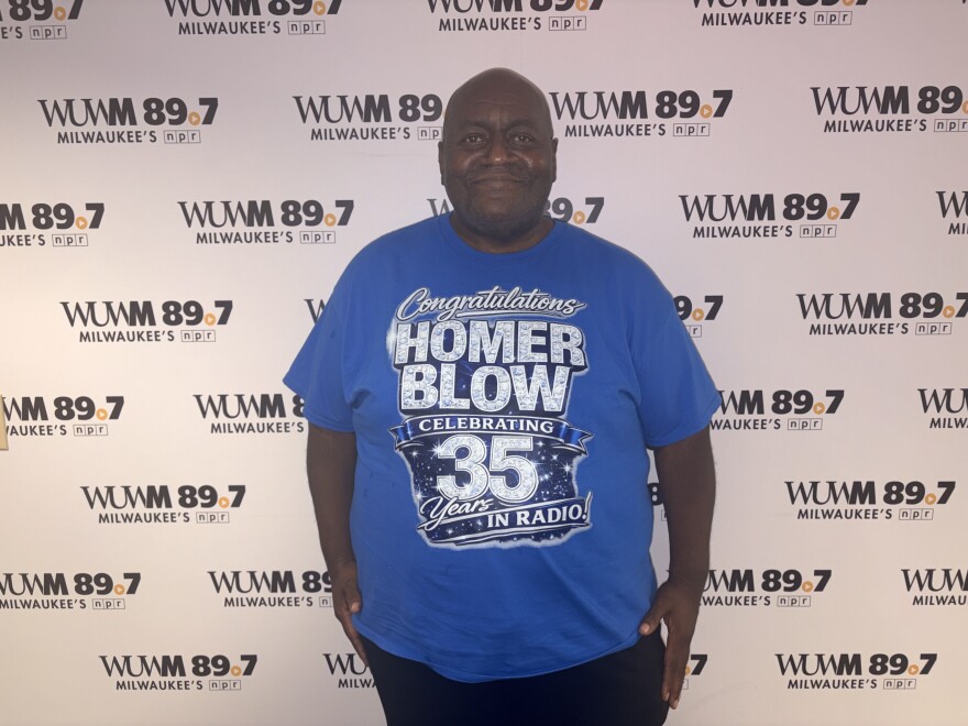 Homer Blow, a long-time radio host in Milwaukee, stands in front of the WUWM 89.7 Milwaukee's NPR photo backdrop wearing a royal blue shirt with the words: "Congratulations Homer Blow Celebrating 35 Years in Radio!"