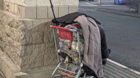  An unhoused person sits outside a grocery store in Longmont, Colorado, with their possessions in a grocery cart.