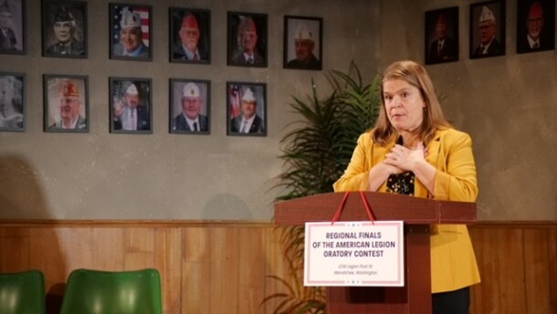 A woman wearing a yellow coat standing in front of a podium with pictures of military servicemen in the background.