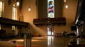 Light pours through the stained glass windows of a church. The pews are gone, and odds and ends are strewn in corners: a ladder, some wooden folding chairs. Center, over a choir balcony, is a tall stained glass window depicting a scene with Jesus. 