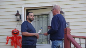 Two men talk in front of the white front door of a white wooden house. To the right of the door stands an orange skeleton with a jack-o-lantern. 