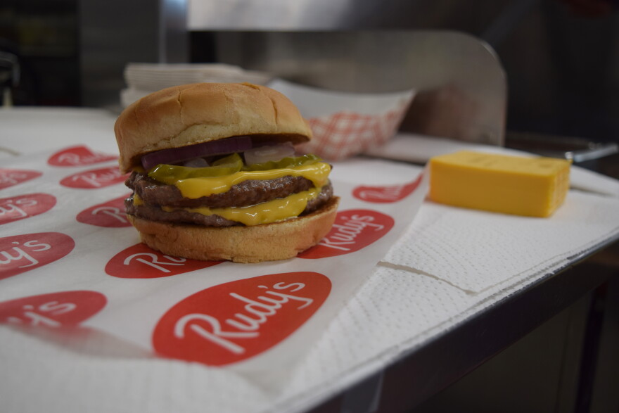 A double cheeseburger with pickles, onions, and cheese sits on branded Rudys paper near a block of cheddar cheese.