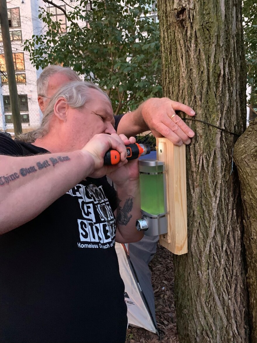 Watchmen of the Streets volunteer Todd Miller installs a hand sanitizer dispenser at a homeless camp east of uptown.