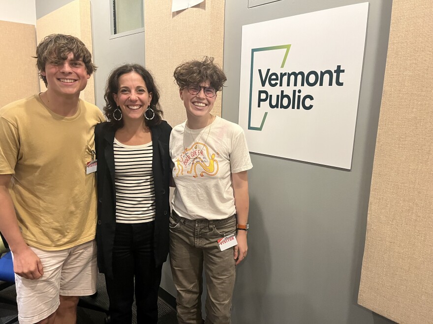 Three people stand together and smile in front of a Vermont Public sign