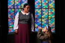 A young woman in a light blue headband kneels with praying hands in front of a woman who looks down at her. They are posed in front of a stained glass window.