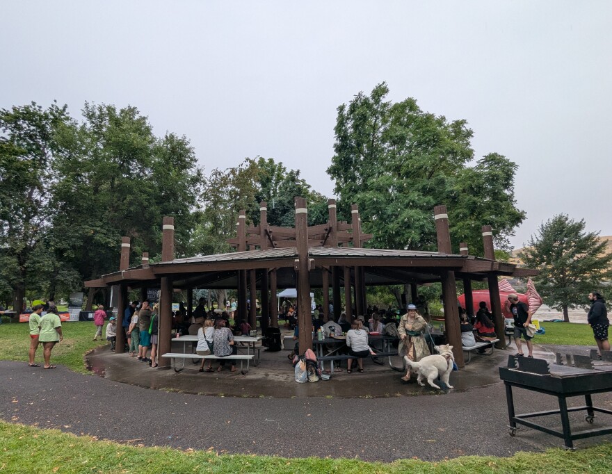 People gather under a gazebo at a park with green grass surrounded by trees.
