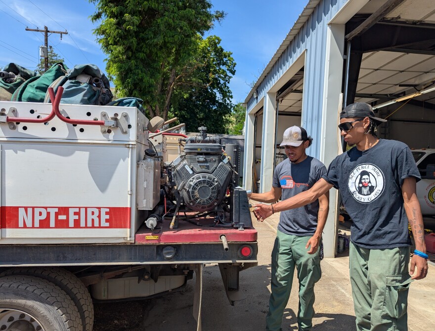 Two young men, wearing baseball hats, t-shirts and green cargo pants, check a pump on the back of a fire truck.