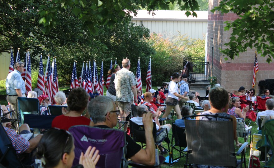 Audience members applaud as a standing veteran a young Scout walk past a row of American flags near the amphitheater stage.