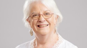 Woman with short white hair and glasses smiles at the camera, wearing a white blouse.
