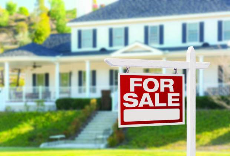 A red FOR SALE sign sits on a lawn in front of a large, white house.