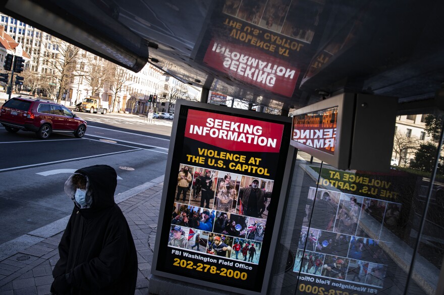 A person wearing a face mask and hood stands next to a city bus stop, where an electronic sign says "Seeking Information: Violence at the U.S. Capitol" above surveillance photographs of suspects.
