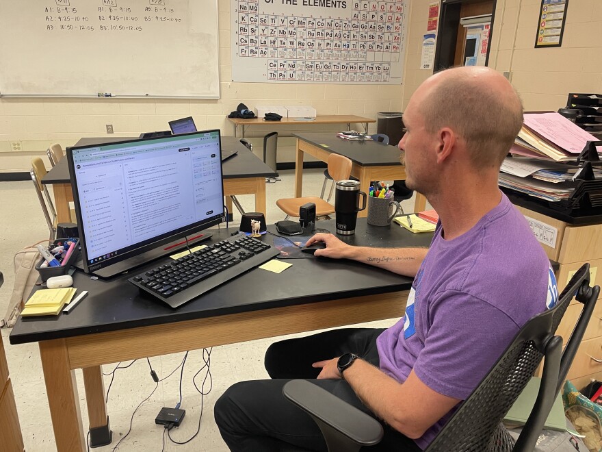 Teacher, Brandt Champion, wearing a purple shirt seats in front of a computer screen looking at it.  