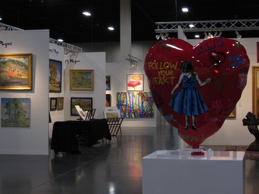 A large sculpted red heart balloon features a young girl in a blue dress holding a red balloon. Graffiti-style text reads "Follow your heart" in yellow. In the background, various painting exhibits are visible.
