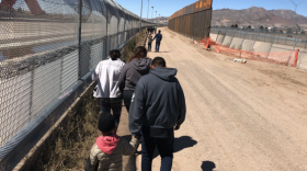 people walking on a dirt road alongside a chainlink fence.