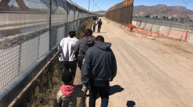 people walking on a dirt road alongside a chainlink fence. 